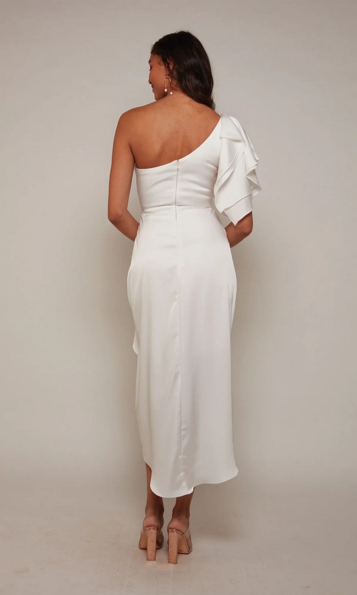 A woman stands facing away in the Alyce 70035 Knee Length Semi-Formal Dress, a white one-shoulder midi with ruffle detail, paired with nude heels against a plain, light background.
